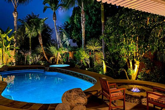 Backyard night view of curved pool with circular gas fire pit, cemented-in-place boulders, curved concrete elevated walkways, dramatic uplighting on a variety of palm trees