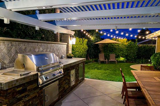 Built-in grill and outdoor food prep area with synthetic turf, string lighting, and alumawood arbor