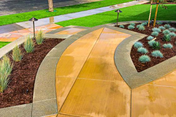 Multicolored poured concrete in a curved shape with clean lines, pathway lights, and evenly spaced grass patches surrounding synthetic turf and masonry