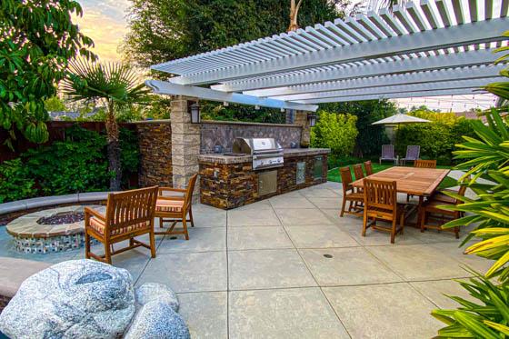 Backyard with boulders, poured concrete, masonry, stone veneer, grill install, alumawood arbor, circular gas firepit, teak deck furniture, and jungle plants