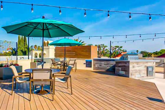 Repurposed multi-family rooftop amenity in Sherman Oaks, California with string lights crossing a blue sky, patio furniture over a wood deck with large potted plants, and an L-shaped grill area