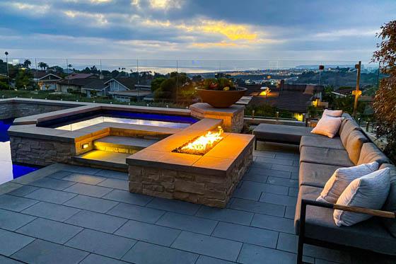 Backyard pool deck featuring inlaid paving stones and a fire pit adjacent to a hot tub with decorative shrubs
