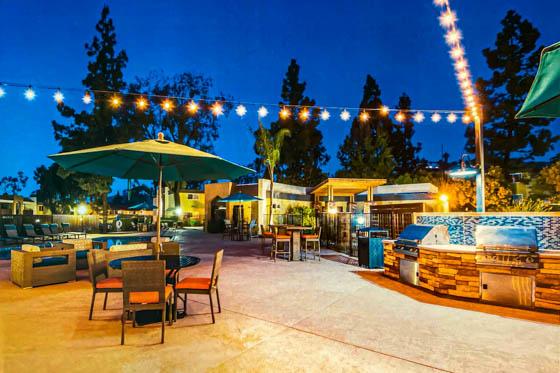 Grill deck at night with festive lighting, stone inlaid backsplash, concrete walking path and tall pine trees in the background near Los Angeles