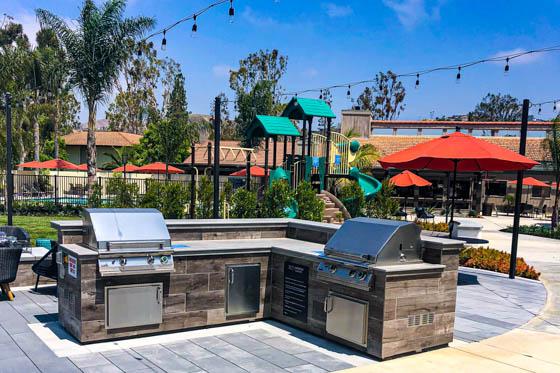 L-shaped outdoor gas grill built into a hardscape with stone veneer and festive lights as part of a congregating space for families