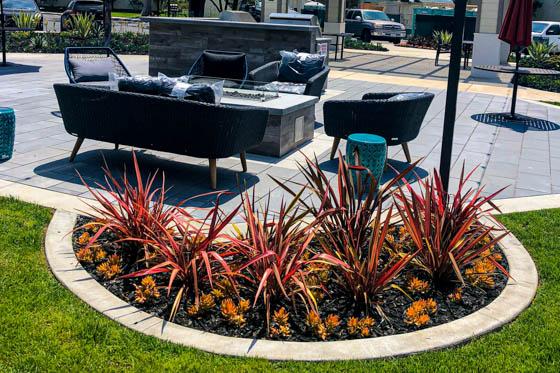 Excellence in poured concrete as demonstrated by a round boundary surrounding a landscaped flowerbed with an elevated fire element, decorative wall, and synthetic turf